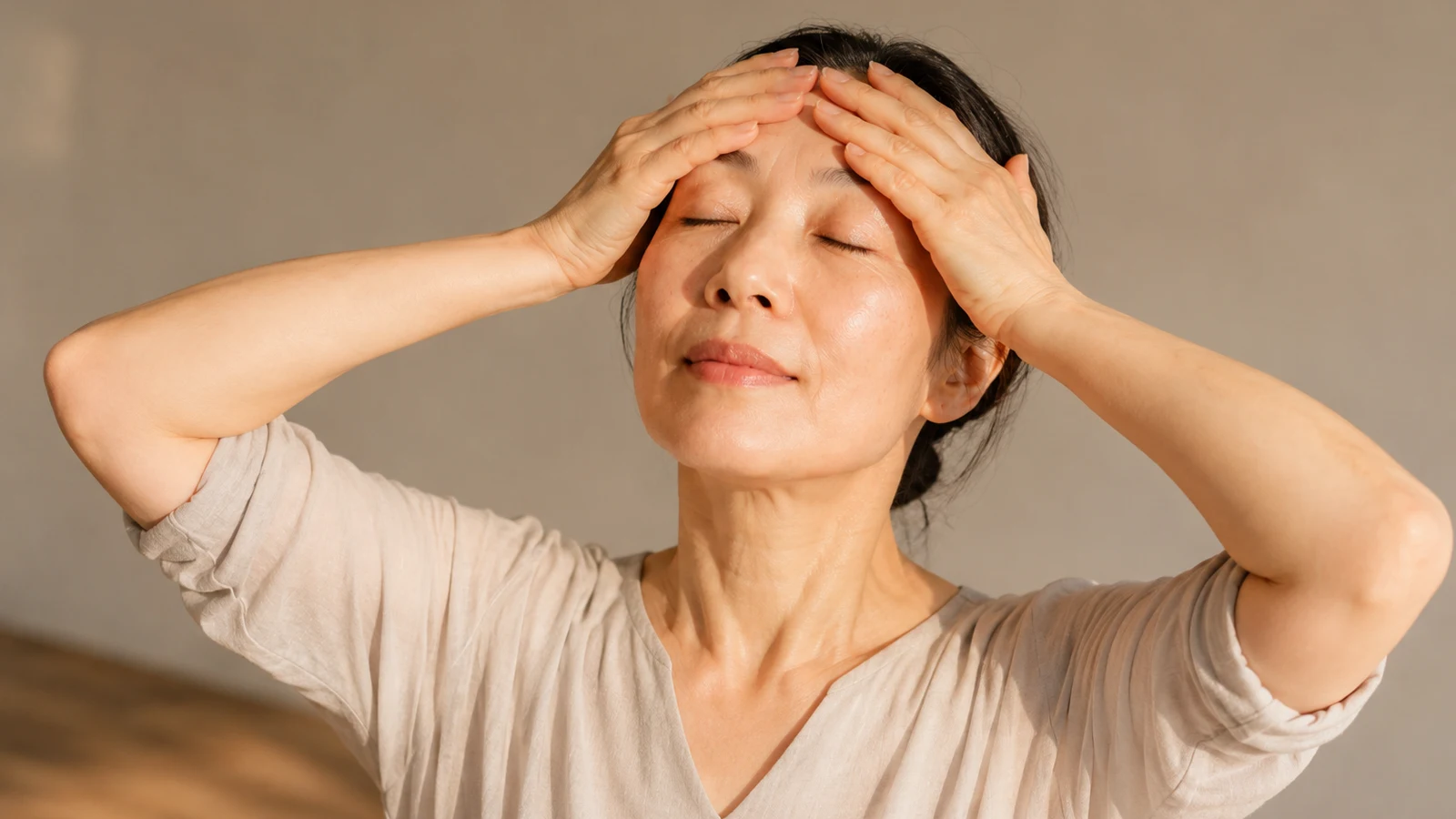 A woman resting both palms gently across her forehead and temples with eyes closed